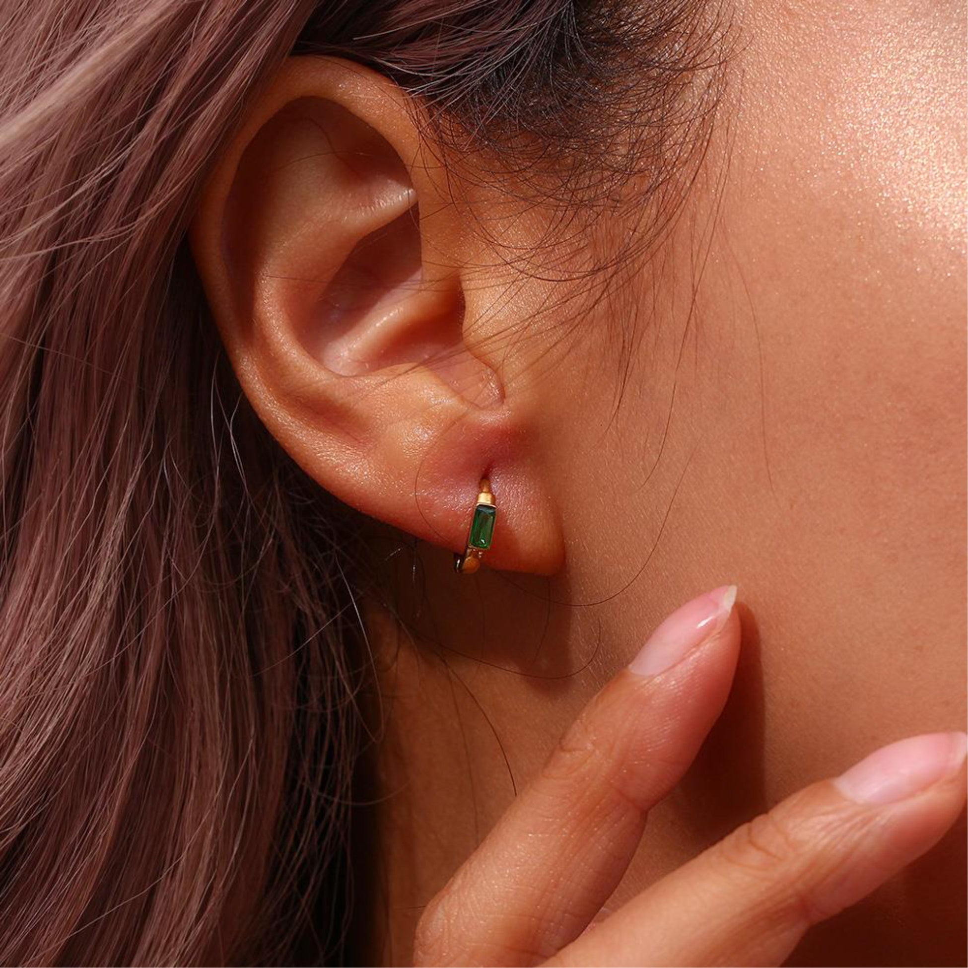 Close-up of an ear wearing a gold hoop earring with green gemstones, with a hand touching the skin.